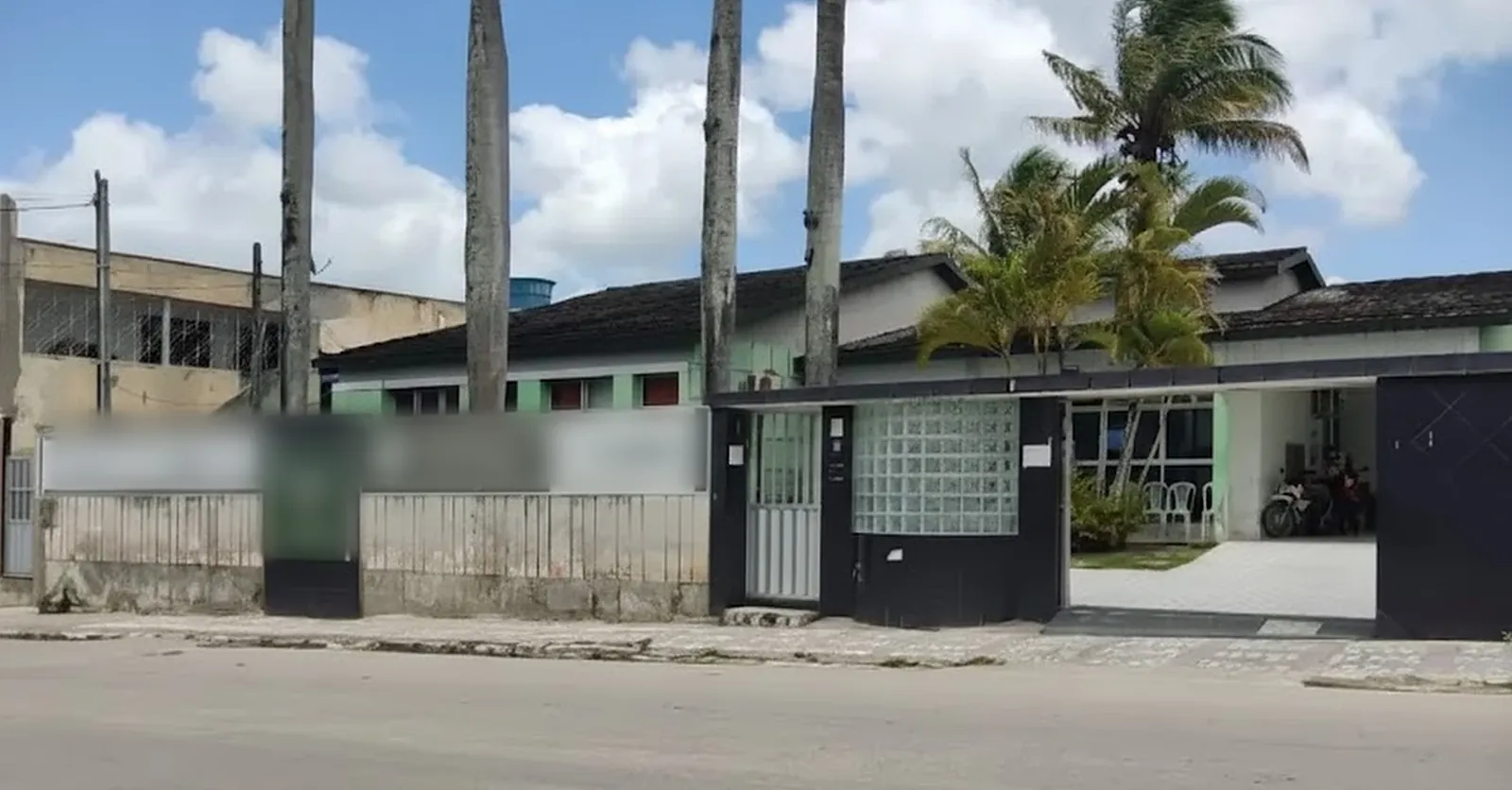 Fachada hoje: muro claro, portão preto, bloco de vidro e casa verde atrás das palmeiras