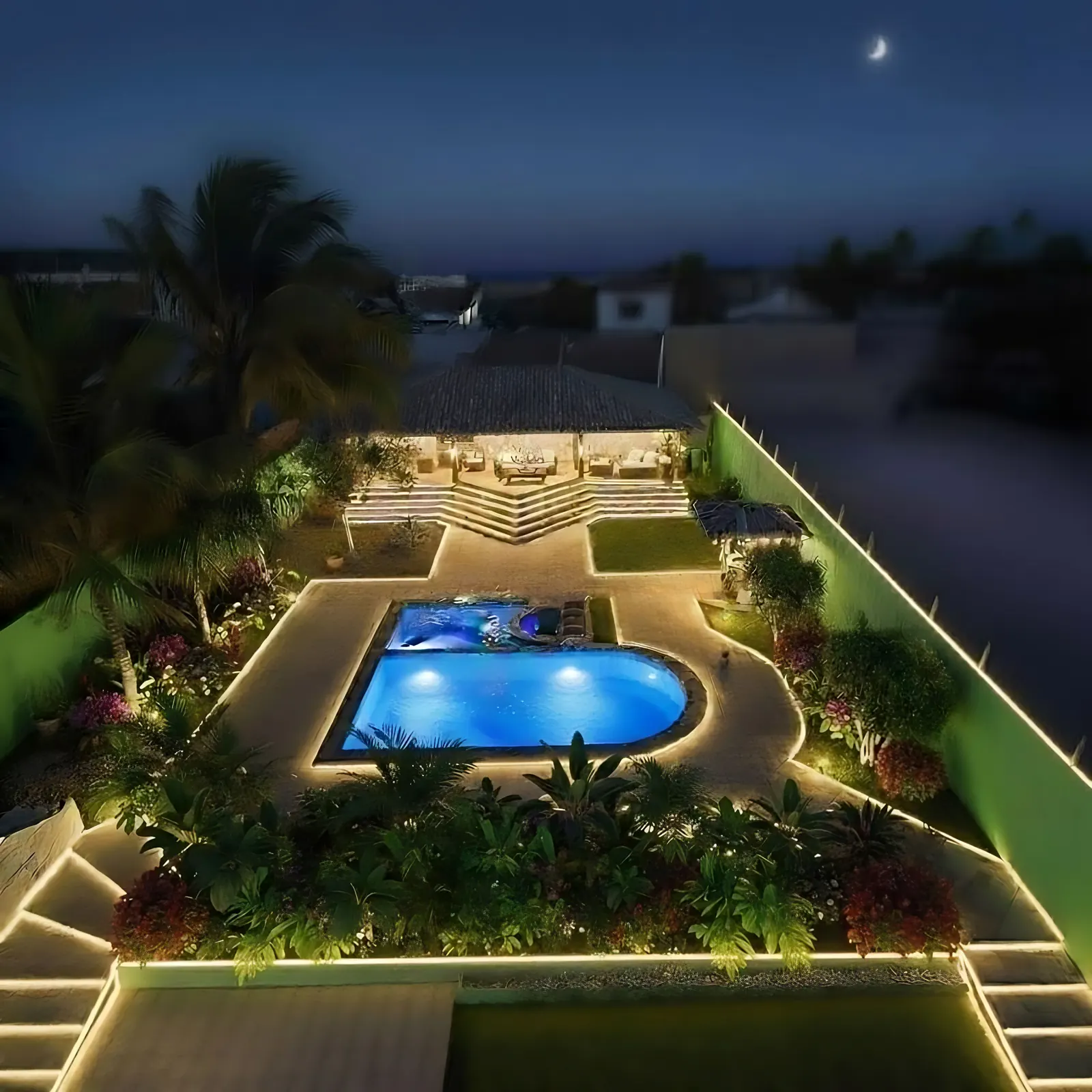 Vista aérea noturna da área de lazer: piscina iluminada, caminhos com luz de contorno, jardim tropical e varanda de convivência ao fundo